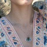 A necklace featuring a hand made thistle-shaped pendant with a green amethyst faceted drop gemstone and micro faceted lavender cubic zirconia gemstones, on a sterling silver bar chain.