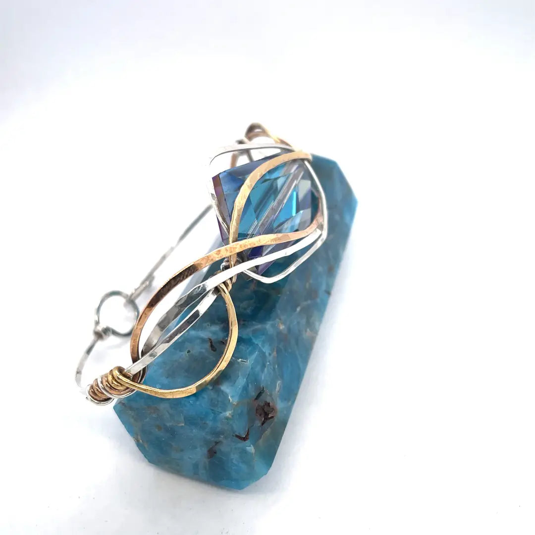 A large blue crystal is nestled within layers of braided hammered sterling silver, brass, and bronze wires to form a handmade statement cuff bracelet