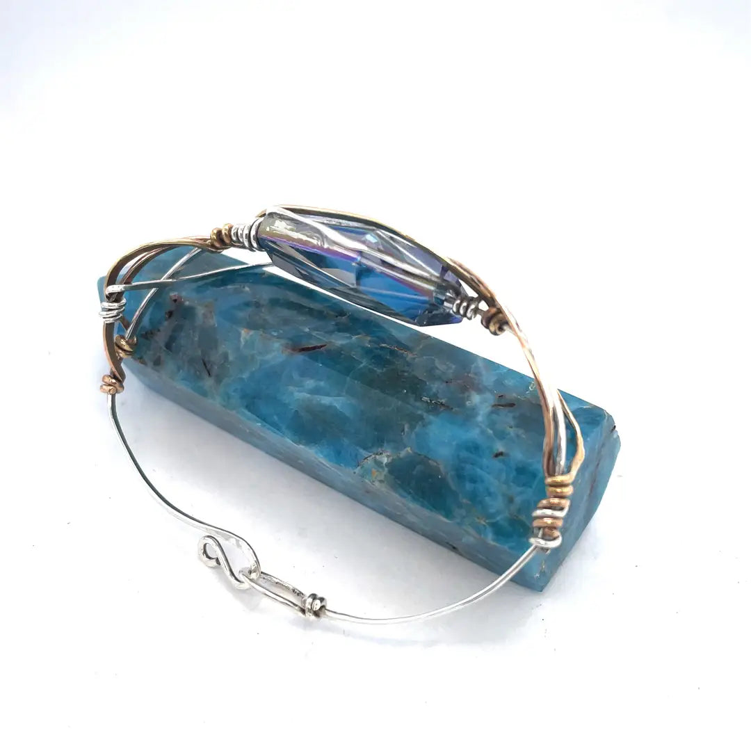 A large blue crystal is nestled within layers of braided hammered sterling silver, brass, and bronze wires to form a handmade statement cuff bracelet