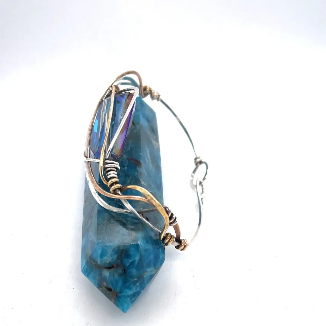 A large blue crystal is nestled within layers of braided hammered sterling silver, brass, and bronze wires to form a handmade statement cuff bracelet