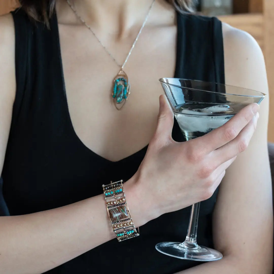 A wide bronze cuff bracelet featuring a combination of striped jasper, fresh water pearls, pyrite, smokey quartz, and turquoise gemstones, strung on sterling silver and hammered bronze.
