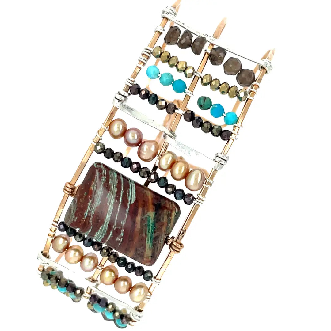 A wide bronze cuff bracelet featuring a combination of striped jasper, fresh water pearls, pyrite, smokey quartz, and turquoise gemstones, strung on sterling silver and hammered bronze.