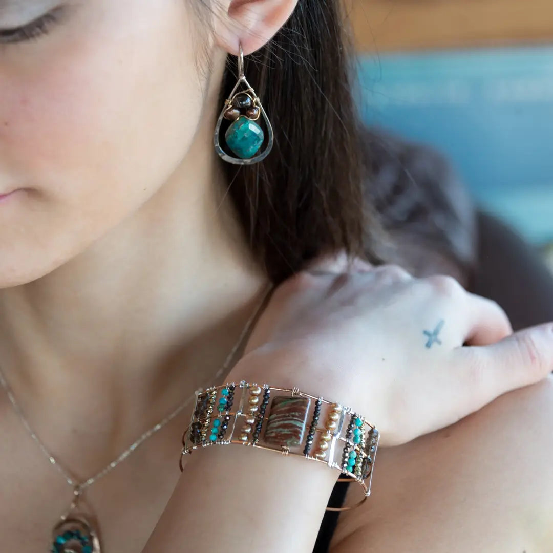 A wide bronze cuff bracelet featuring a combination of striped jasper, fresh water pearls, pyrite, smokey quartz, and turquoise gemstones, strung on sterling silver and hammered bronze.