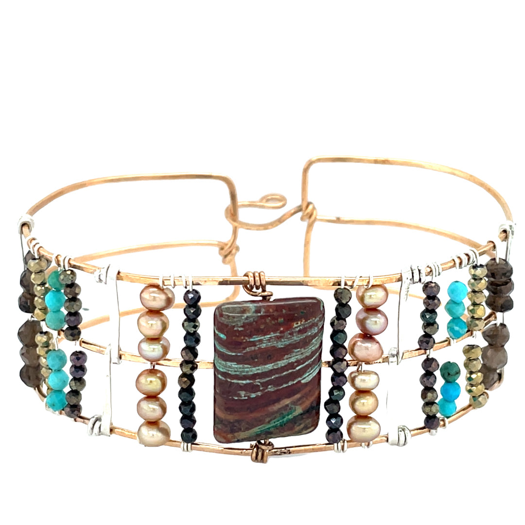A wide bronze cuff bracelet featuring a combination of striped jasper, fresh water pearls, pyrite, smokey quartz, and turquoise gemstones, strung on sterling silver and hammered bronze.