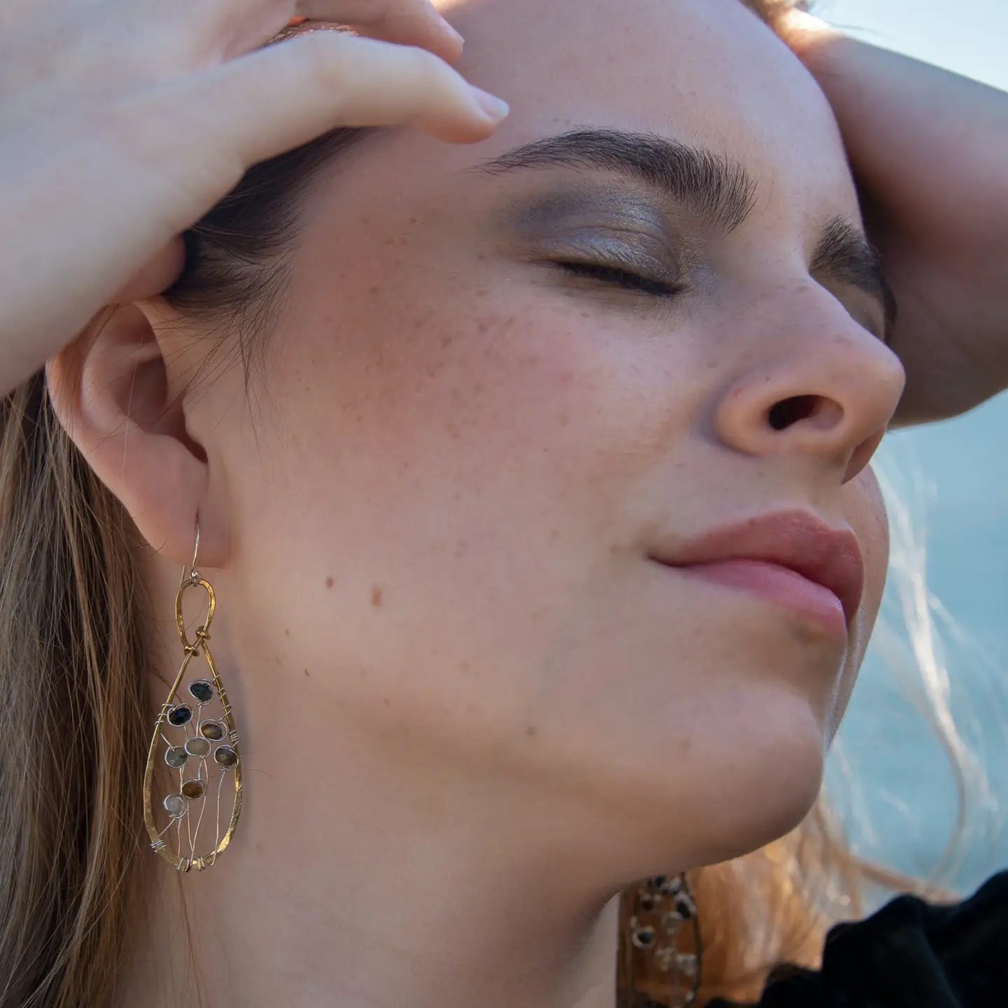 A pair of brass infinity shaped earrings featuring multicolored cat's eye faceted gemstones  and sterling silver details. Natural shades of yellow, blue, and green