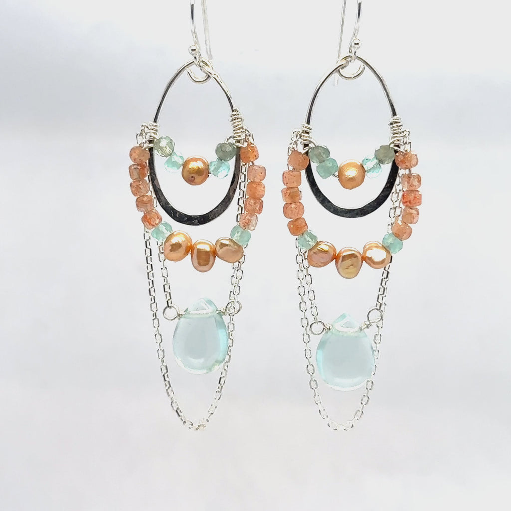 A pair of chandelier earrings featuring silver ovals, with sunstone, chalcedony, fresh water pearl, and apatite gemstones, and a single aqua quartz drop. 
